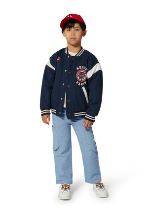 Veste universitaire bleu marine avec des accents blancs et un logo de tigre, associée à un pantalon cargo bleu clair et des baskets blanches ornées d'un motif de tigre. Casquette rouge.