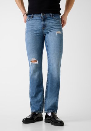 Persona che indossa jeans a gamba dritta di colore azzurro chiaro con strappi sul ginocchio sinistro e sulla coscia destra, abbinati a mocassini neri e calzini bianchi.