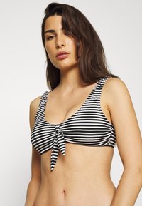 Black and white striped bikini top with a knotted front detail and wide straps, featuring a smooth texture and a fitted silhouette.