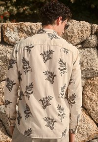 Beige košile s tmavým květinovým vzorem kapradí, dlouhými rukávy, zadním yokes a knoflíkovými manžetami. Lehce sepraná látka s jemným, texturovaným vzhledem.