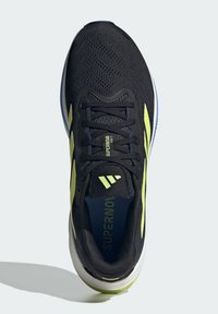 Zapatilla de correr negra con parte superior de malla, acentos amarillos, diseño con cordones, cuello acolchado y suela texturada para tracción. Marca visible en la lengua.