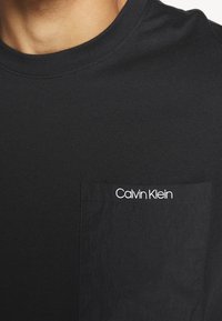 Černé bavlněné tričko s kulatým výstřihem a náprsní kapsou z kontrastní látky. Na kapse je bílé logo "Calvin Klein".