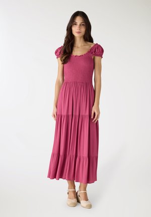 Donna con capelli lunghi e scuri che indossa un vestito rosa con maniche a sbuffo arricciate e scarpe beige con zeppa e cinturino alla caviglia su sfondo bianco.