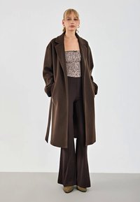 Cappotto oversize marrone in lana con colletto a punte, indossato sopra un top floreale aderente e pantaloni scuri a vita alta. Stivali alla caviglia verdi completano il look.