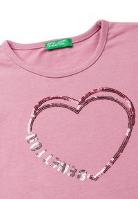Růžové bavlněné tričko s designem srdce vyrobeným ze stříbrných a růžových flitrů, s nápisem značky "BENETTON" pod ním také z flitrů.