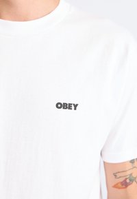 White cotton t-shirt featuring a small black "OBEY" logo on the left chest area, with a classic crew neckline and short sleeves.