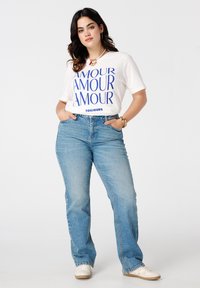Witte t-shirt met blauwe tekst "AMOUR" en "TOUJOURS" onderaan, gecombineerd met blauwe flared jeans en witte sneakers. Gouden accessoires zichtbaar.