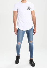 Man som bär en vit t-shirt med svart logotyp och rivna smala blåjeans tillsammans med svarta sneakers, stående mot vit bakgrund.