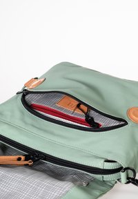 Helle grüne Tasche mit strukturiertem Finish, ausgestattet mit schwarzen Reißverschlüssen und hellbraunen Lederakzenten. Das Innere enthält ein rotes Fach und ein Gittermuster-Futter.