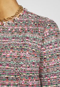 Kolorowy teksturowany top z plecionym wzorem z różowych, zielonych i czarnych nici; postrzępiony dekolt; złoty łańcuszek jako ozdoba.