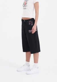 Shorts de mezclilla negra con un corte holgado, con bolsillos de estampado floral y detalles de texto bordado en la parte frontal. Llevados con zapatillas blancas.