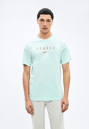 Muž v svetlomodrom tričku s nápisom „FRANCÚZSKO“ a oranžovým logom Nike, v svetlosivých nohaviciach, stojaci na bielom pozadí.