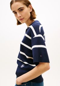 Μπλε ναυτικό και άσπρη ριγέ πλεκτή μπλούζα με κοντά μανίκια, που διαθέτει λαιμόκοψη τύπου crew και κεντημένο λογότυπο. Μπλε τζιν παντελόνι ορατό στη μέση.