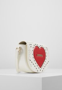 Bílá kožená kabelka přes rameno s výrazným červeným srdcem, perforovanými detaily a zlatým logem na přední straně.