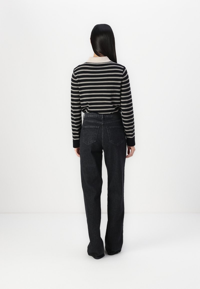 Femme aux longs cheveux foncés portant un haut à manches longues rayé noir et blanc et un jean taille haute foncé, debout face à un mur blanc.