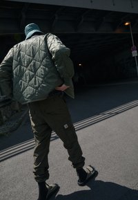 Kurtka pikowana w kolorze oliwkowej zieleni, spodnie cargo i sandały. Materiał o fakturze, luźny krój. Scena pod wiaduktem, środowisko miejskie, obecne cienie.