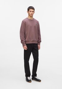 Sudadera morada con puños acanalados, que presenta un pequeño logo, combinada con pantalones negros y zapatillas negras con cordones blancos y suela de goma.