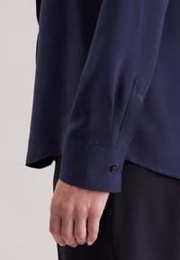 Käsi lepää sivulla, yllään tummansininen pitkähihainen paita, jossa on napitettu kalvosin, ja mustat housut, yksinkertaista vaaleaa taustaa vasten.