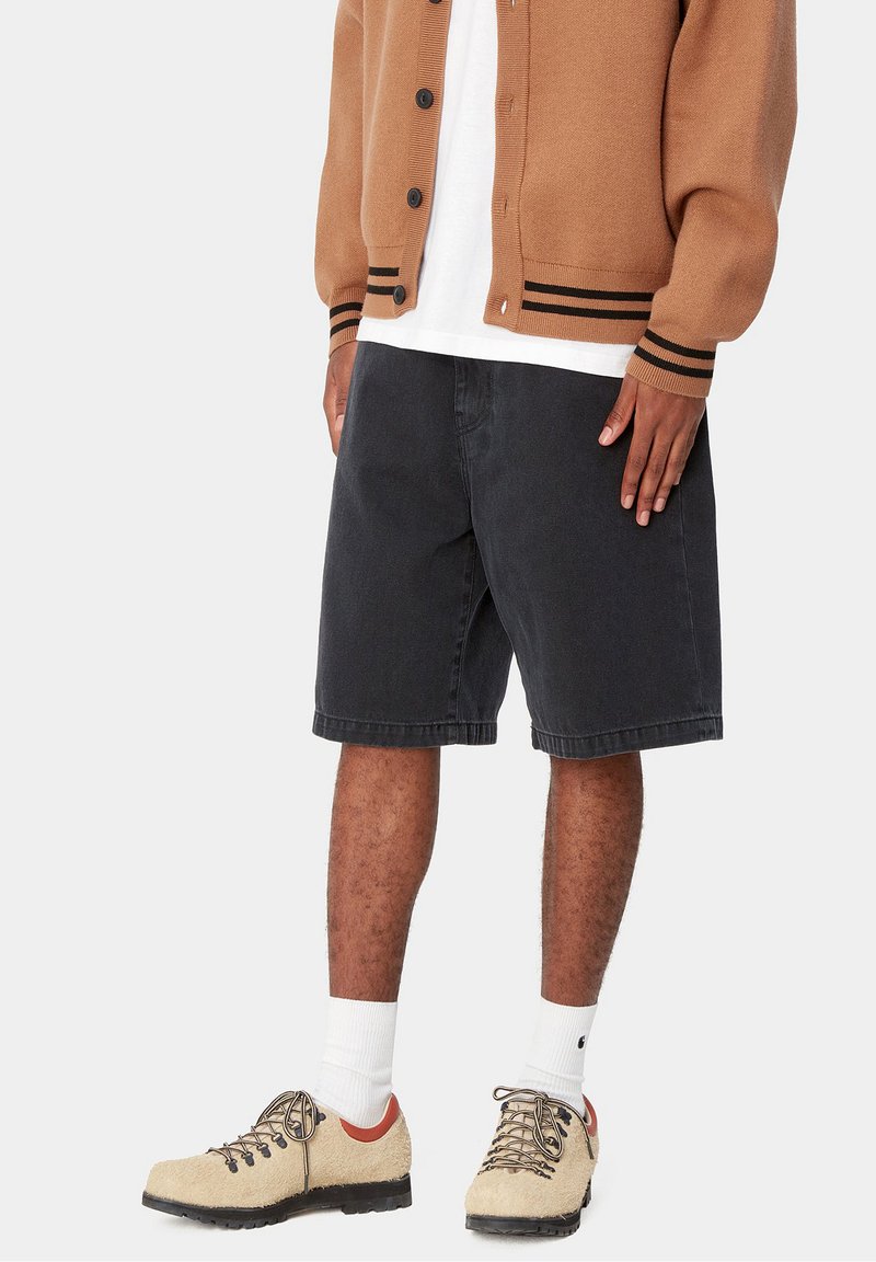 Underkroppen af en person iført sorte denimshorts, beige teksturerede sneakers med sorte såler, hvide sokker og en brun cardigan over en hvid skjorte.