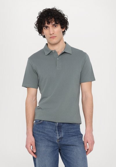 Short-sleeve polo shirt in a muted green color, featuring a classic collar, two-button placket, and a textured material. Worn with blue jeans.