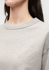 Persona que lleva una sudadera gris claro con cuello redondo acanalado y letras bordadas en blanco que forman la palabra "CLOSED" en el pecho.