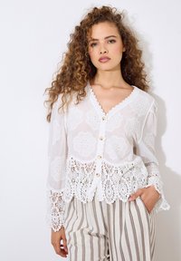 Weißes Hemdblusenshirt mit floraler Stickerei und Spitzeneinsatz am Saum und den Ärmeln. Leichter Stoff und ein V-Ausschnitt ergänzen gestreifte Hosen.