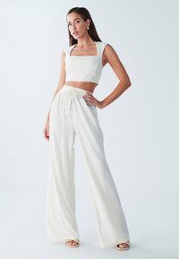 Bílý zkrácený top se čtvercovým výst�řihem a širokými kalhotami. Materiál má texturovaný, žebrovaný design. Modelka má na sobě sandály v neutrálních tónech.