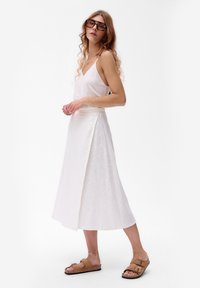 Witte linnen jurk met een flared silhouet en een zijsplit. Gecombineerd met bruine sandalen. Model heeft krullend haar en draagt grote zonnebrillen.