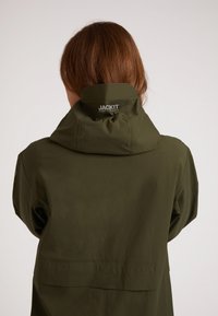 Olivgrön vattentät jacka med justerbar huva, med en logga på kragen och en slät matt finish. Strukturerad, praktisk design.