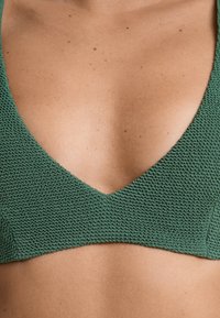 Grön stickad bralette med djup V-ringning, som har en texturerad typ av tyg och släta kanter. Designen är minimalistisk utan synlig hårdvara.