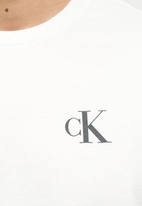 Λευκό πουκάμισο με μαύρο λογότυπο "cK" τυπωμένο στο στήθος κοντά στον λαιμό.