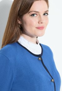 Kungablå cardigan i strukturerad stickad kvalitet, med marinblå kant och två runda knappar framtill, lager på lager över en vit blus.