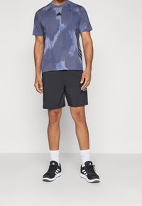 Blå och grå tie-dye atletisk t-shirt med mesh-textur och svarta ränder, parat med svarta shorts och svarta sportskor.