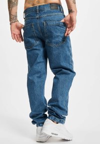 Jeans de mezclilla azul gastados con un ajuste relajado, bolsillos traseros y costuras visibles. Combinados con zapatillas blancas.