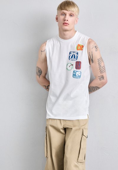 Tricou alb fără mâneci, cu applique grafice colorate. Asortat cu pantaloni cargo bej, evidențiind o croială lejeră și buzunare.