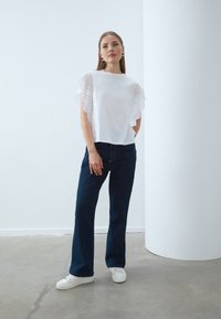 Bílý top s krajkovými volánkovými rukávy, zkombinovaný s tmavě modrými rozšířenými džínami a bílými teniskami. Neutrální pozadí a jednoduchý design.