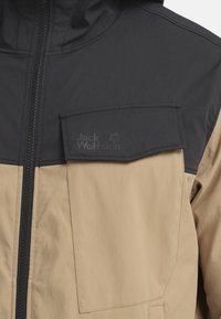 Wasserabweisende Jacke mit einem zweifarbigen Design, bestehend aus einem schwarzen oberen Teil und einem khakifarbenen unteren Teil, Reißverschluss und geprägt Logo auf der Brusttasche.