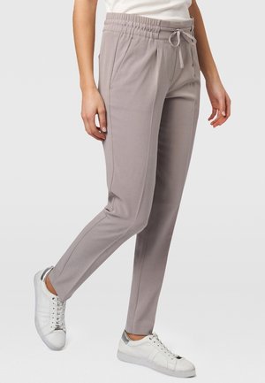 Person trägt hellgraue, tapered Hosen mit elastischem Bund und Kordelzug, kombiniert mit weißen Sneakers mit silbernen Details.