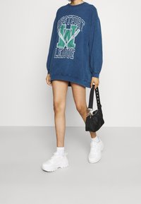 Person som bär en överstor blå sweatshirt med texten "Westfort League", vita sneakers, vita strumpor och håller en liten svart handväska.