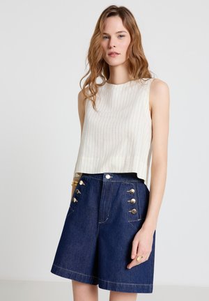 Giovane donna con capelli mossi che indossa una canottiera bianca a righe e pantaloncini di denim scuro a vita alta con bottoni dorati.