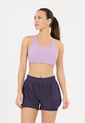 Vrouw met donker haar in een paardenstaart, die een lichtpaarse sportbeha en donkere sportshorts draagt, staande tegen een witte achtergrond.