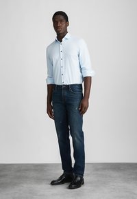 Ανοιχτό μπλε πουκαμίσα με μακρύ μανίκι, σκούρα μπλε τζιν με φθορές και μαύρες παντόφλες, στέκεται σε γκρι φόντο.