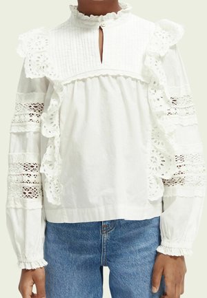 Weiße Langarmbluse mit Spitzenpaneelen, Rüschen und kleinem Frontschlitz, kombiniert mit blauen Jeans bei einer dunkelhäutigen Person.