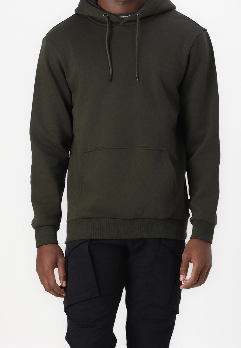 Homme portant un sweat à capuche vert foncé avec poche avant et un pantalon cargo noir, debout devant un fond blanc uni.