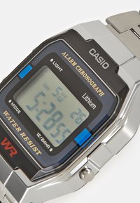 Montre numérique avec un cadran rectangulaire, fond noir, bracelet en acier inoxydable argenté et accent bleu sur le côté. Affichage des heures et de la date.