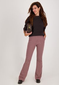 Vrouw die staat in gestructureerde wijde broek, zwarte top, met een bruine handtas, lang bruin haar en gouden creolen.