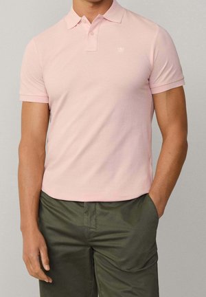 Mann trägt ein hellrosa kurzärmliges Poloshirt, das in olivgrüne Freizeithosen gesteckt ist, steht mit einer Hand in der Tasche.