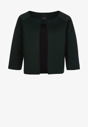 Mujer caminando con una chaqueta corta de color verde oscuro, camiseta beige, vaqueros ajustados oscuros y botines negros de tacón alto puntiagudos, llevando un gran bolso negro.