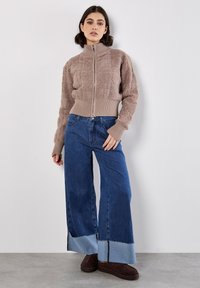 Suéter beige de felpa con cremallera y dobladillo acanalado, combinado con pantalones de mezclilla azul de pierna ancha que tienen un acento de dobladillo de mezclilla clara y zapatos marrones.
