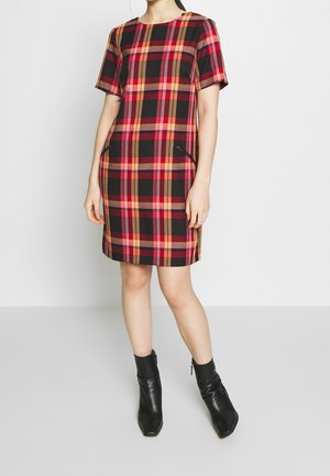 Kobieta w sukience w kratę z krótkim rękawem, sięgającej do kolan, w kolorach czerwonym, różowym i brązowym, z kieszeniami na zamek, w połączeniu z czarnymi botkami za kostkę.